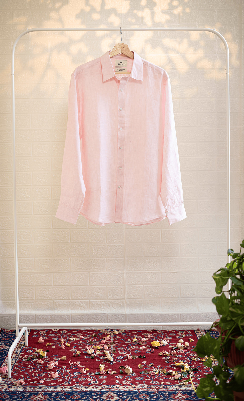 Blush / Pink shirt on a hanger against a neutral wall with a patterned rug and plant in the foreground.
