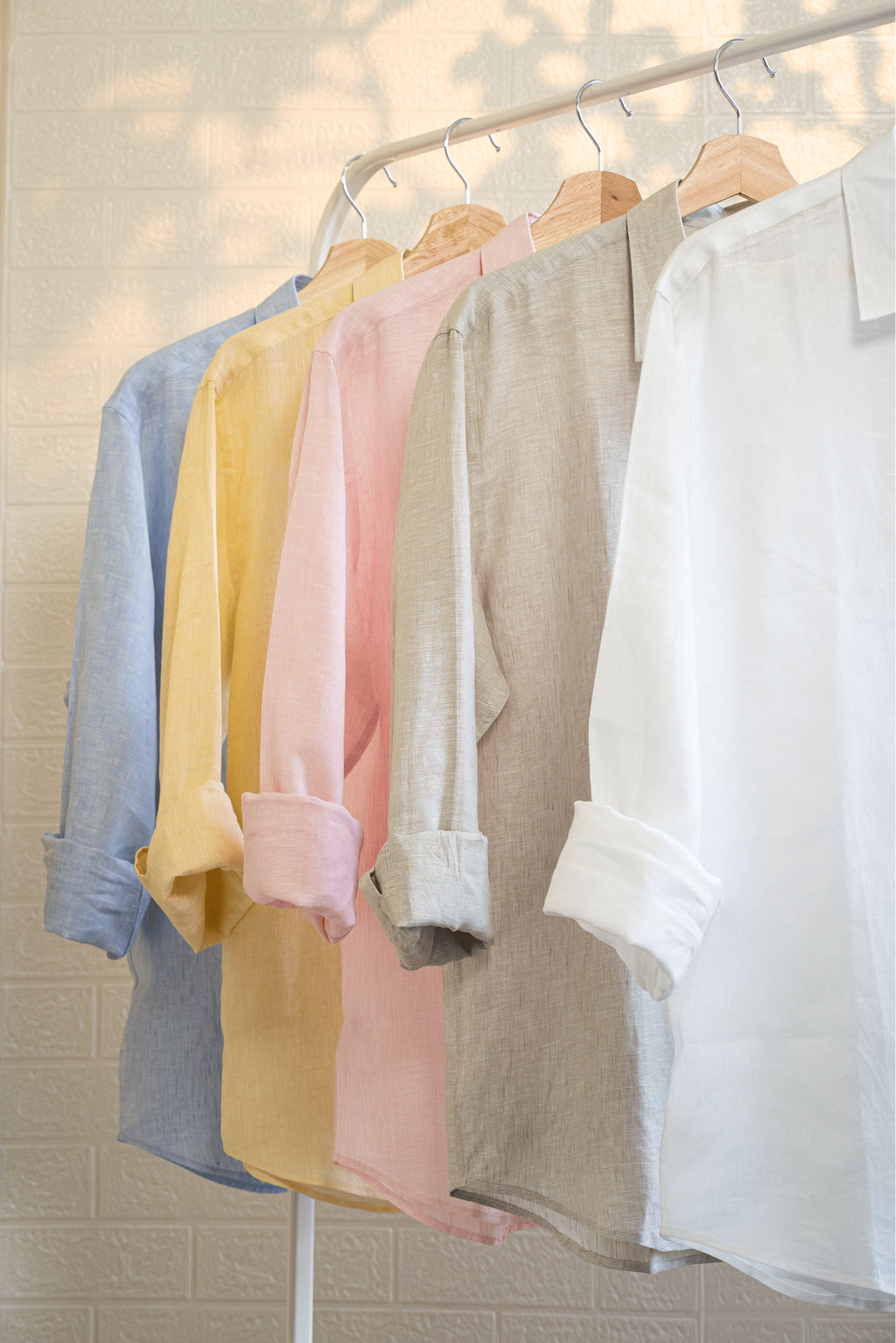 Set of pastel-colored shirts hanging on a rack against a neutral background. Sand  shirt is second from the right